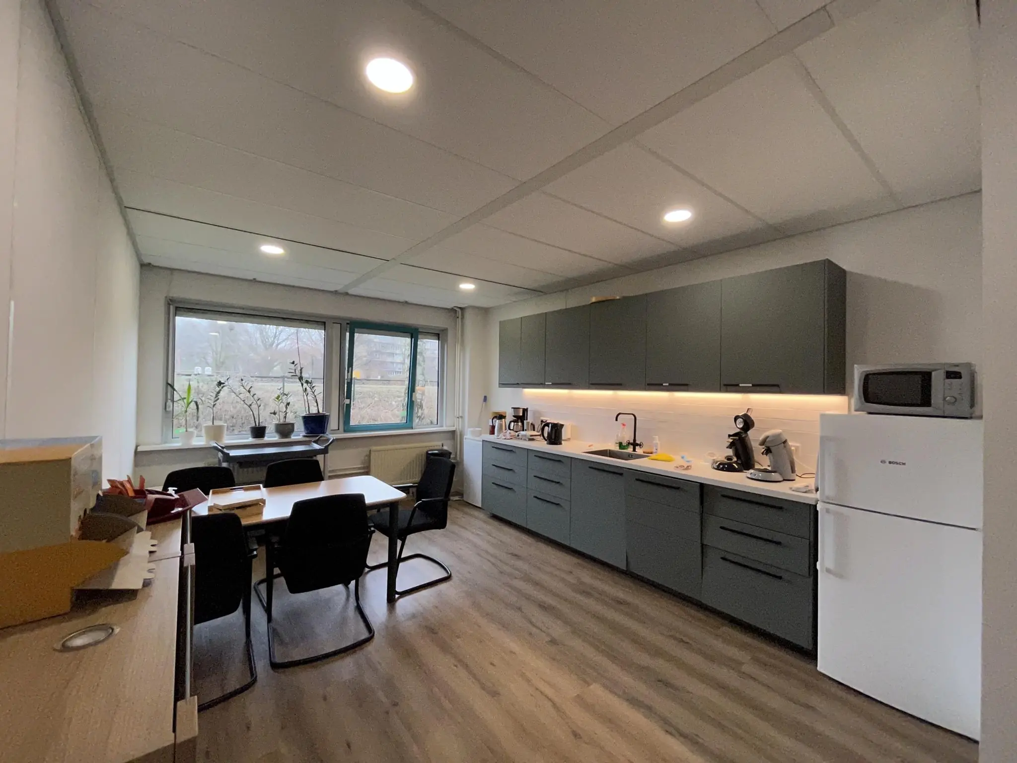 Moderne kantoorkeuken aan de Christiaan Krammlaan met donkergrijze keukenkasten, koelkast, koffiemachines en een vergadertafel bij het raam.